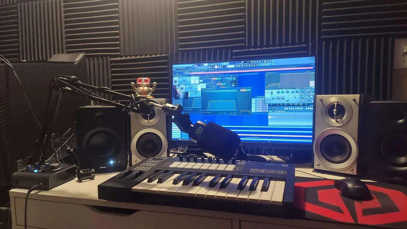 A home studio: upright piano against the far wall, an audio interface and monitors on the desk, guitars on wall hooks.